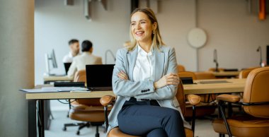 Gri takım elbiseli kendine güvenen bir kadın modern bir ofiste bir anlık eğlencenin tadını çıkarır. Arka planda işbirliği yapan insanlarla çevrili bir şekilde otururken gülümsüyor..
