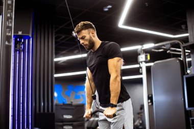 Odaklanmış bir adam şık bir spor salonunda kablo triseps uzantıları yapıyor, gelişmiş fitness ekipmanları ve canlı ışıklandırma ile çevrili..