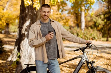 Genç bir adam sıcak bir içki tutuyor ve güzel bir sonbahar parkında bisikletine yaslanırken gülümsüyor. Altın yapraklar sarmış onu.
