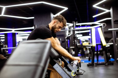 Bir adam parlak neon ışıklarla çevrili çağdaş bir spor salonunda çalışıyor. Bir makinenin üzerine oturmuş, egzersiz rutinine odaklanmış, kendini spora adamış..