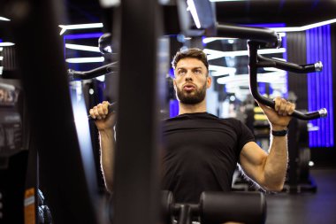 Odaklanmış bir yüz ifadesi olan bir adam iyi aydınlatılmış bir spor salonunda ağırlık kaldırıyor. Ekipman ve ışıklandırma, spor salonundaki deneyimini geliştirmek için canlı bir atmosfer yaratıyor..