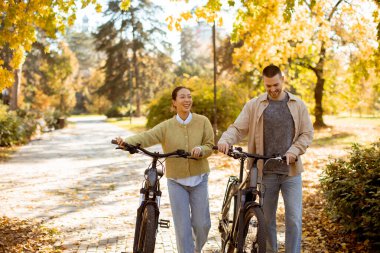 İki arkadaş bisikletlerle yan yana yürüyor, gülümsüyor ve renkli yapraklarla dolu canlı sonbahar manzarasının tadını çıkarıyorlar..