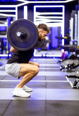 Bir adam ağır bir halterle, şık ve canlı bir spor salonunda, idman sırasında kendini adamış ve güçlü bir şekilde çömeliyor..