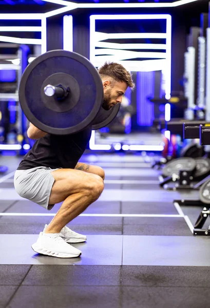 Bir adam ağır bir halterle, şık ve canlı bir spor salonunda, idman sırasında kendini adamış ve güçlü bir şekilde çömeliyor..