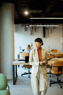 Profesyonel kıyafetli bir kadın, şık bir çalışma alanında yürürken bir not defteri taşır. Meslektaşlar masalarda otururlar, tartışmalara katılırlar veya bireysel olarak çalışırlar..