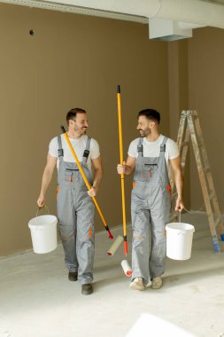 Gri tulumlu iki işçi ellerinde boya kovaları ve bigudilerle yeni boyanmış bir odadan geçiyorlar. Birlikte gülüyorlar ve işlerinin tadını çıkarıyorlar. Arka planda bir merdiven görünüyor..