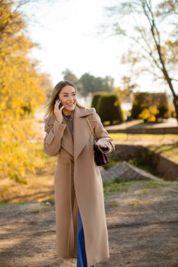 Şık ceketli genç bir kadın sonbahar yapraklarıyla süslenmiş bir parkta geziniyor. Telefonla konuşurken gülümsüyor, dışarıda güzel bir günün tadını çıkarıyor..