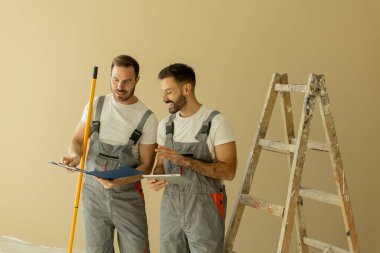 Gri tulumlu iki erkek işçi yenileme planlarını gözden geçiriyorlar. Gülümsüyorlar ve yakınlarda bir merdiven ve boya aletleriyle sohbet ediyorlar. Oda tadilat için hazırlanıyor..