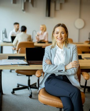 A woman sitting in a stylish chair smiles confidently in a bright office. Colleagues are chatting and working at desks nearby, showcasing teamwork and a vibrant work environment.