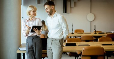 İki meslektaş, iş birlikçi bir atmosferle çevrili modern bir ofis ortamında, bir tablet hakkında konuşur ve analiz eder..