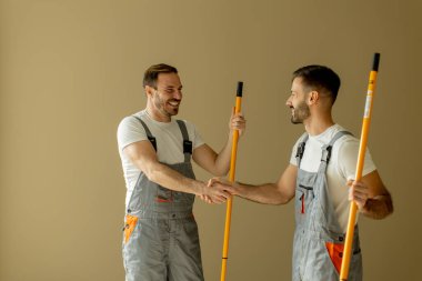 Gri tulumlu iki adam açık renkli bir odada duruyor. El sıkışıyorlar ve uzun boyama direklerini tutuyorlar. Proje için hazırlar. Atmosfer dostane ve profesyonel görünüyor..