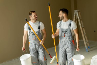 İki adam boş bir odada boya rulosu ve kova tutarken gülümsüyor ve sohbet ediyorlar. Duvarları boyamaya hazırlanıyorlar, takım çalışmalarını açık renkli bir ortamda sergiliyorlar..