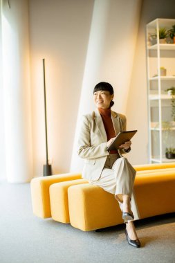 Açık renkli takım elbiseli bir iş kadını elinde tabletle sarı bir bankta oturuyor. Modern ofisin keyifli atmosferinin tadını çıkarırken gülümsüyor..