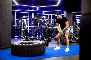 Bir adam enerji dolu bir şekilde çekiçle büyük bir lastiğe vuruyor. Pürüzsüz, modern bir spor salonunda..