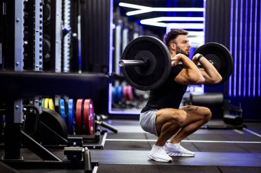 Çağdaş bir spor salonunda canlı ışıklandırmalı halter çömelmesi yapan bir adam. Etrafı çeşitli ağırlık plakaları ve ekipmanlarla çevrili olan antrenmanına dikkatle odaklanır..