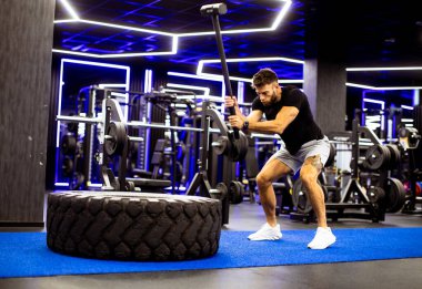 Bir adam güçlü bir şekilde balyozla egzersiz yapar, gösterişli bir spor salonunda büyük bir lastiğe vurur. Parlak ışıklar ve spor aletleri arka planı dolduruyor ve enerjik atmosfere ekliyor..