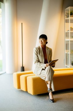 Hafif takım elbiseli kendine güvenen bir kadın şık bir ofis ortamında sarı banklarda oturur. Elinde bir tablet tutuyor, parlak doğal ışık altında pencereden dışarı derin derin bakıyor..