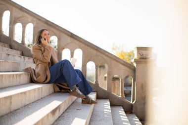 Genç bir kadın parkta taş basamaklarda oturuyor, sıcak bir içkiyi yudumlarken telefonla konuşuyor. Rahat bir ceket ve rahat bir kot pantolon giyer. Doğayla iç içe güneşli bir sonbahar gününün tadını çıkarır..