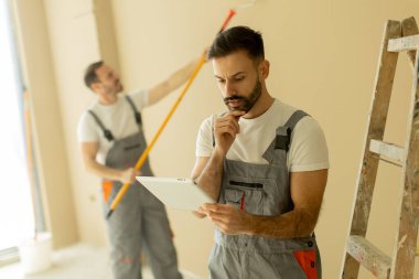 İki adam bir odada birlikte çalışıyor. Bir adam bir silindirle duvarı boyuyor, diğeri de tablete bakıyor, muhtemelen proje detaylarını gözden geçiriyor. Görevlerine odaklanmış görünüyorlar..