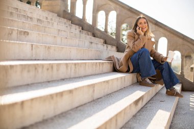Bir kadın taş basamaklarda rahatça oturur, telefonda sohbet ederken kahvesini yudumlar. Güneş ışıl ışıl parlıyor, rahat duruşunu ve şık kıyafetini vurguluyor..