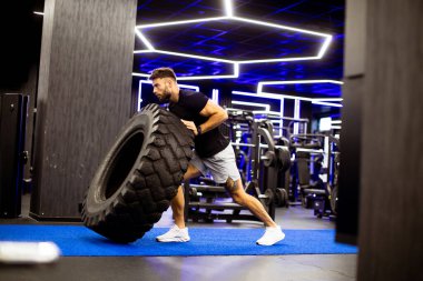Modern bir spor salonunda bir adam mavi bir paspasın üzerinde büyük bir lastik itiyor. Siyah bir gömlek ve şort giyerek antrenmanına odaklanmış. Spor salonunda onu çevreleyen parlak ışık ve egzersiz ekipmanları var..