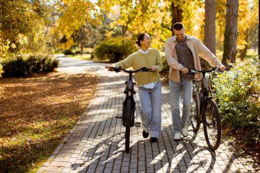 Koreli bir kadın ve beyaz bir adam altın yapraklarla dolu güneşli bir parkta bisikletleriyle yürüyüp sohbet ediyor ve gülümsüyor..