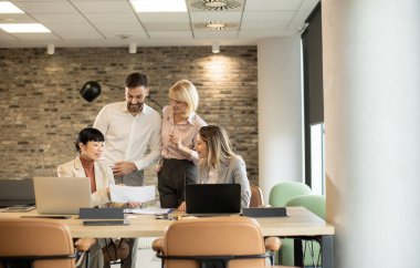 Aydınlık ve modern bir ofiste, dört takım üyesi verimli bir toplantı yapıyor. Dizüstü bilgisayarları ve not defterleri etrafa saçılmış büyük bir masada otururken bir belgeyi tartışıyorlar..