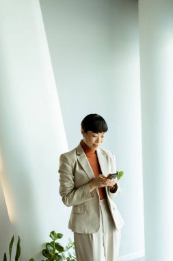 Hafif takım elbiseli bir kadın şık bir ofis alanında duruyor ve akıllı telefonuna odaklanıyor. Yeşil bitkiler ve doğal ışık rahatlatıcı bir atmosfer yaratır..