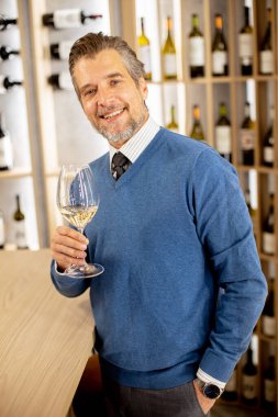 Bir adam barda dikilirken bir bardak beyaz şarap tutuyor. Onun arkasında çeşitli şarap şişeleriyle dolu raflar var. O bir kazak ve kravat giydi..