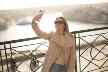 A woman stands by a railing in Malta, taking a selfie with her smartphone. The sun sets over the water, creating a warm glow. She enjoys the moment and captures the view.