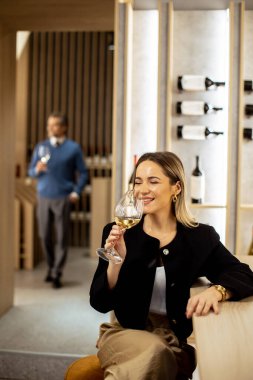 Barda elinde bir bardak beyaz şarap tutan bir kadın gülümsüyor. Arka planda bir adam yürüyor, o da içki taşıyor. Sergide ahşap dekor ve şarap şişeleri yer alıyor..