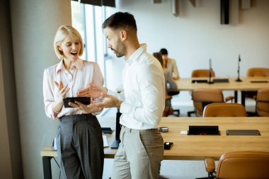 İki meslektaşı bir tableti incelerken projeleri hakkında canlı bir tartışmaya giriştiler. Ofisin arka planda çalışan insanlarla dolu modern bir tasarımı var..