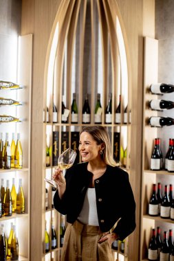 Bir kadın şarap dükkanında elinde bir bardak şarapla durup gülümsüyor. Arkasında çeşitli şarap şişeleriyle dolu raflar var. Mağaza ahşap özellikli modern bir tasarıma sahip..