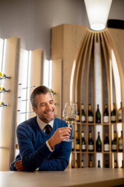 Adamın biri elinde beyaz şarap bardağıyla barda oturuyor. İçkiye bakarken gülümsüyor. Arkasında, raflarda şarap şişeleri var. Mekanın sıcak bir atmosferi var..