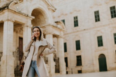 Malta 'da telefonunu kullanan bir kadın var. O, tarihi bir binanın yakınında. Gündüz oldu ve güneş parlıyor. Kadın rahatlamış ve konuşmasına dalmış görünüyor..