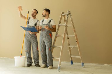 İki işçi merdiven ve aletlerle bir odada duruyor. Planları tartışıyor ve duvarları boyamaya hazırlanıyorlar. Yanlarında bir kova boya var..
