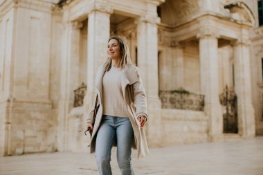 Bir kadın Malta 'da tarihi bir bölgeden geçiyor. Sıradan giyinmiş ve mutlu görünüyor. Güneş ışığı çevreyi incelerken etrafında sıcak bir ortam yaratıyor..