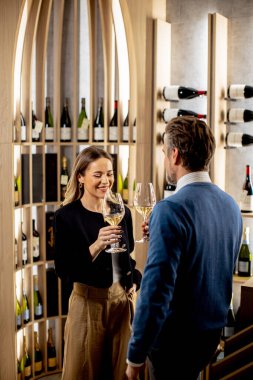 Şarap mahzeninde şarap kadehiyle kadeh kaldıran iki kişi. Kurulumda şarap şişeleriyle dolu ahşap raflar yer alıyor. Bir anlık neşeyi paylaşıyor gibi görünüyorlar, gülümsüyorlar ve birbirlerine bağlanıyorlar..