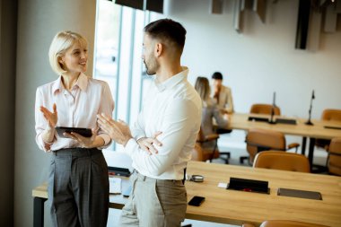 İki iş adamı parlak ve modern bir ofiste dururken bir projeyi tartışıyorlar. Diğer meslektaşlar arka planda çalışırken görülebilir, işbirlikçi bir atmosfer yaratırlar..