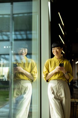 Cam bir duvarın yanında elinde akıllı bir telefonla duran bir kadın gülümsüyor..