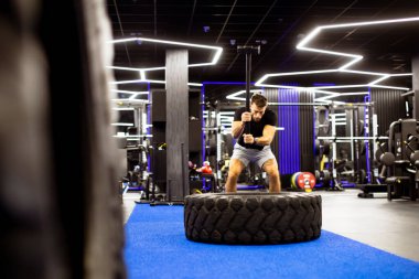 Çağdaş bir spor salonunda bir kişi lastik çevirme egzersizi yapıyor. Uzay modern ekipmanlar ve parlak ışıklandırmalar içeriyor, fitness meraklıları için enerjik bir atmosfer yaratıyor..