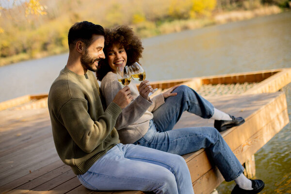 Two friends sit on a wooden deck by a calm lake, laughing and toasting with glasses of wine under the warm autumn sun.