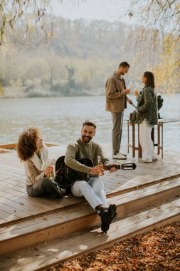 Bir grup arkadaş sakin bir göl kenarında dinlenir, güneşli bir sonbahar gününde müzik dinlerken içkilerini ve kahkahalarını paylaşırlar..