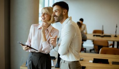 İki meslektaş fikirleri tartışırken gülümser ve modern ofis ortamında bir tablete bakarlar. Atmosfer canlı ve yaratıcı. Arka planda diğer ekip üyeleri var..
