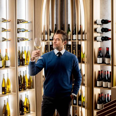 Bir adam kendine güvenerek elinde bir kadeh şarap tutarak, çeşitli şişelerle dolu şık bir şaraphanede aromasının tadını çıkarıyor..