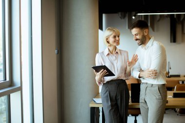 Modern bir ofiste iki iş arkadaşı duruyor. Bir tanesi büyük bir pencerenin yanında konuşurken bir tablet tutuyor ve birbirlerine gülümsüyor. Ayar parlak ve meşgul görünüyor..