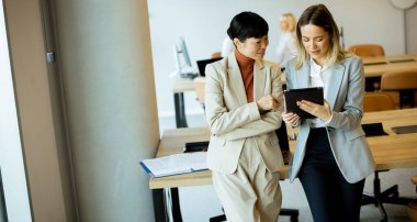 İş kıyafetli iki kadın, geniş ve modern bir ofiste tableti incelerken sohbet ediyorlar. Ayarda geniş pencereler ve çağdaş mobilyalar yer alıyor.