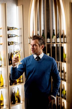 İyi giyimli bir adam, elinde bir bardak şarap tutarak, şarap mahzeninin rengini ve berraklığını takdir eder..