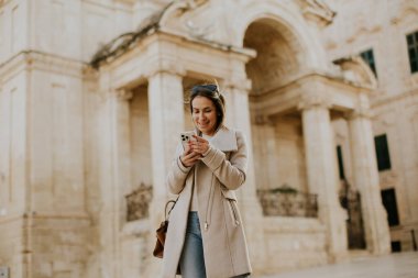 Malta 'da bir kadın akıllı telefonuna bakarken sokakta yürüyor. Taş mimarisi olan eski bir binanın yanında. Gündüz vakti ve manzara bölgedeki yerel yaşamı gösteriyor..