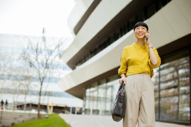 Sarı bluzlu ve açık renk pantolonlu bir kadın telefonla konuşurken gülümsüyor. Bir çanta taşıyor ve güneşli bir günde geniş pencereli, gösterişli, modern bir binanın yanında yürüyor..